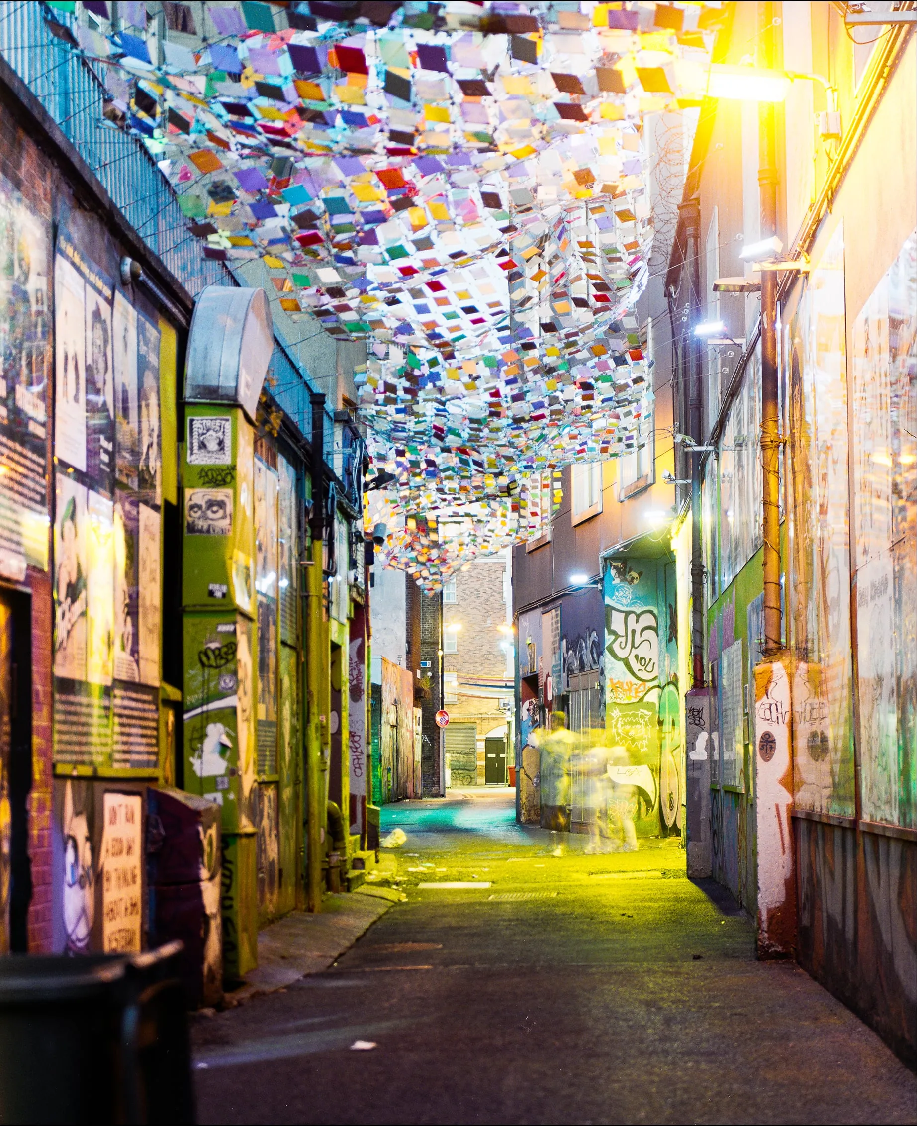 Graffitied Dublin alley at night under a canopy of colored panels