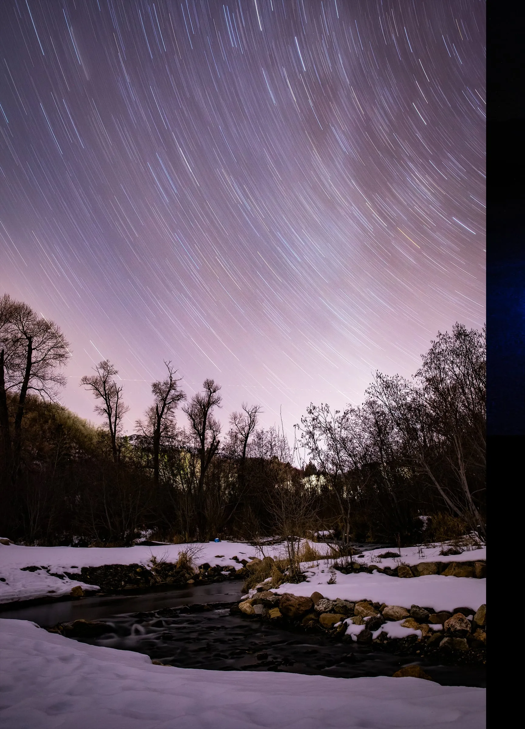 Star trails streaking over a snowy creek lined with bare trees